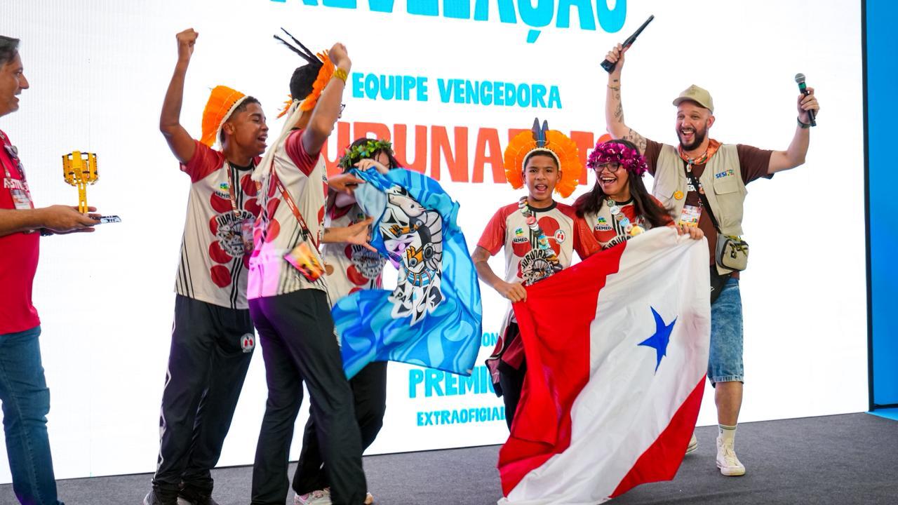 Estudantes indígenas comemoram no palco durante uma premiação de robótica. O grupo veste camisetas da equipe Jurunabots e alguns utilizam adornos tradicionais indígenas. Eles celebram com os braços levantados, segurando uma bandeira do Pará e um pano azul com o símbolo da equipe. Ao fundo, um telão exibe a palavra “Premiação” e o nome da equipe vencedora. Um apresentador segura um microfone ao lado do grupo, enquanto os estudantes demonstram alegria e entusiasmo pela conquista.