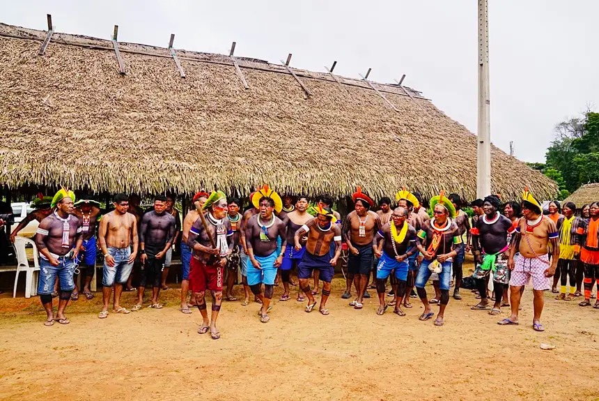 Indígenas um ao lado do outro em uma paisagem de cão de terra e telhado de palha ao fundo