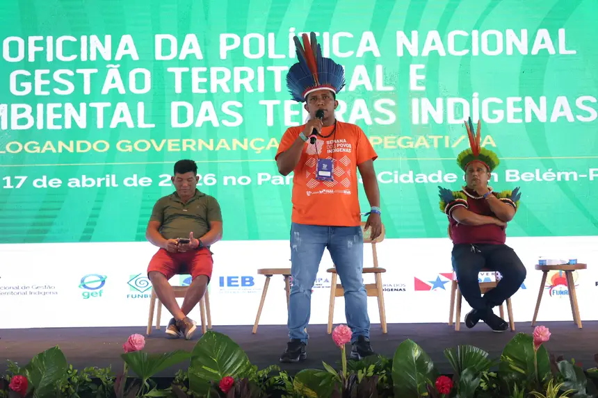 Homem indígena, usando cocar com penas coloridas e camiseta laranja, fala ao microfone em um palco durante um evento. Ao fundo, um telão exibe o título da oficina sobre a Política Nacional de Gestão Territorial e Ambiental de Terras Indígenas, com menção a Belém e à data do encontro. Dois outros homens, também com vestimentas casuais e um deles com cocar, estão sentados em cadeiras atrás do palestrante. A frente do palco é decorada com plantas ornamentais.