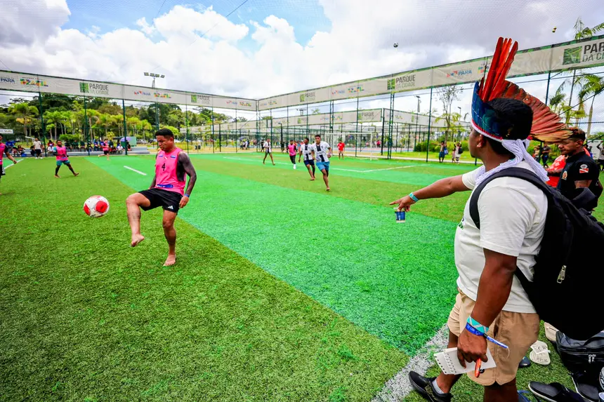Jovens participam de uma partida de futebol em um campo de grama sintética ao ar livre. Em primeiro plano, um jogador descalço, vestindo colete rosa, domina a bola com o pé. À direita, um homem observa e aponta para o jogo, usando um cocar com penas vermelhas e uma mochila nas costas. Ao fundo, outros jogadores se movimentam em quadras cercadas por grades, com áreas esportivas e vegetação ao redor, sob um céu parcialmente nublado.