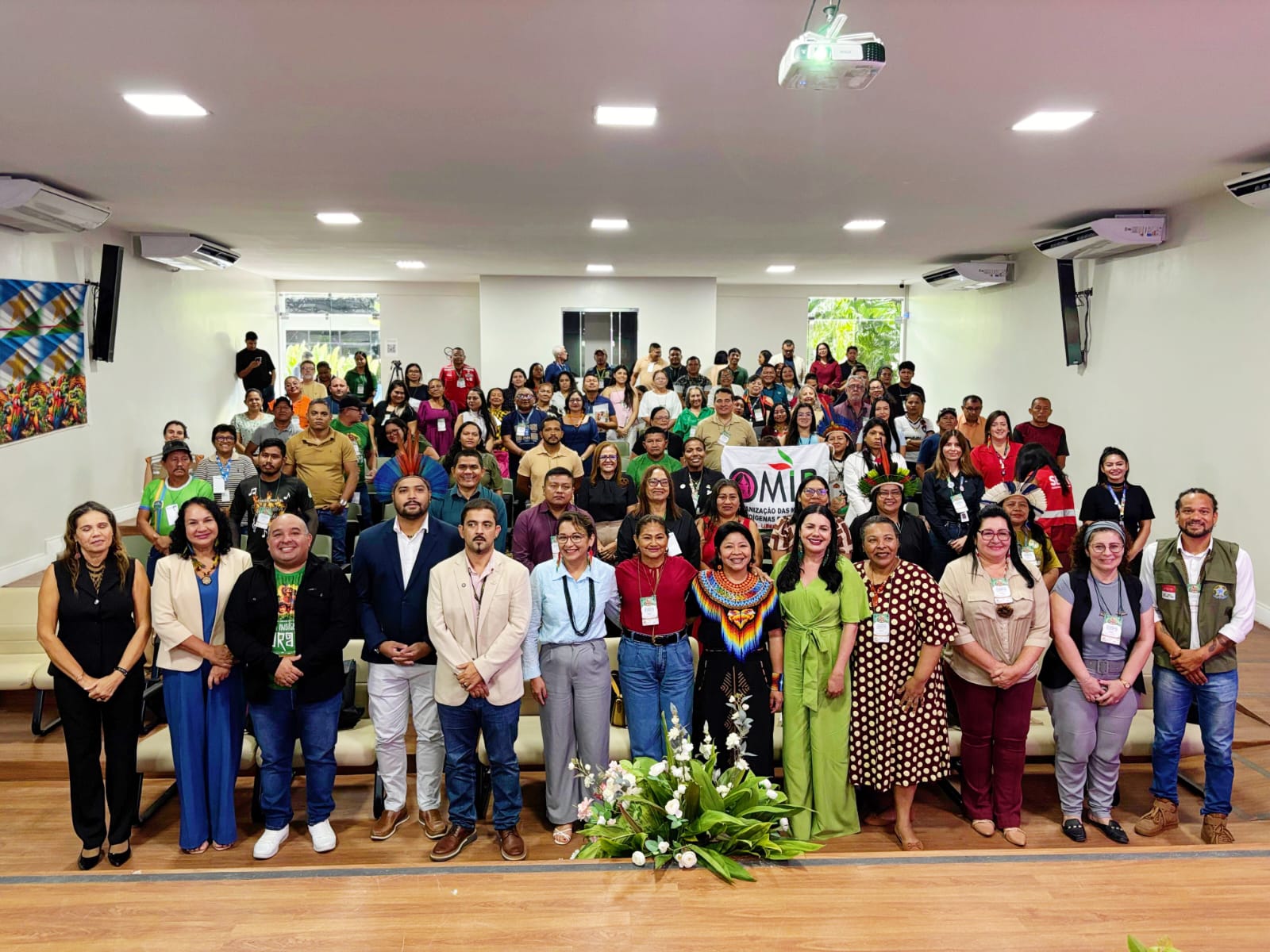 Grupo grande de pessoas posando para foto em um auditório, com o secretário adjunto entre os participantes. Algumas pessoas usam trajes tradicionais indígenas, enquanto outras estão com roupas formais ou casuais. Ao fundo, há um banner do evento, e na frente, um arranjo de flores no chão.