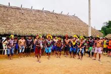 Indígenas um ao lado do outro em uma paisagem de cão de terra e telhado de palha ao fundo