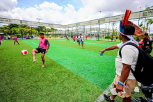 Jovens participam de uma partida de futebol em um campo de grama sintética ao ar livre. Em primeiro plano, um jogador descalço, vestindo colete rosa, domina a bola com o pé. À direita, um homem observa e aponta para o jogo, usando um cocar com penas vermelhas e uma mochila nas costas. Ao fundo, outros jogadores se movimentam em quadras cercadas por grades, com áreas esportivas e vegetação ao redor, sob um céu parcialmente nublado.