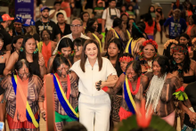 A governadora Hana de roupa clara caminha ao centro de um grupo de mulheres indígenas em um espaço interno movimentado. Elas estão de mãos dadas, algumas com pinturas corporais, colares e adornos tradicionais, formando uma roda ou cortejo. A mulher ao centro sorri enquanto acompanha o grupo. Ao fundo, há uma multidão observando, com pessoas em pé e algumas registrando o momento. O ambiente é iluminado e cheio de cores, destacando a diversidade de vestimentas e expressões culturais.