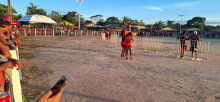 Cena em uma arena de terra ao ar livre, com público ao fundo atrás de cercas e tendas montadas. No centro, duas pessoas com roupas vermelhas participam de uma disputa corporal, em contato direto, enquanto outras observam ao redor.